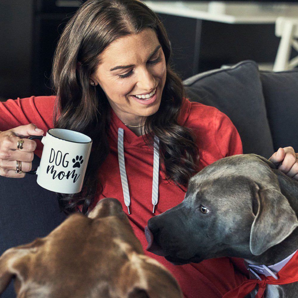 woman's soccer player Alex Morgan with dog mom mug sitting on couch petting dogs
